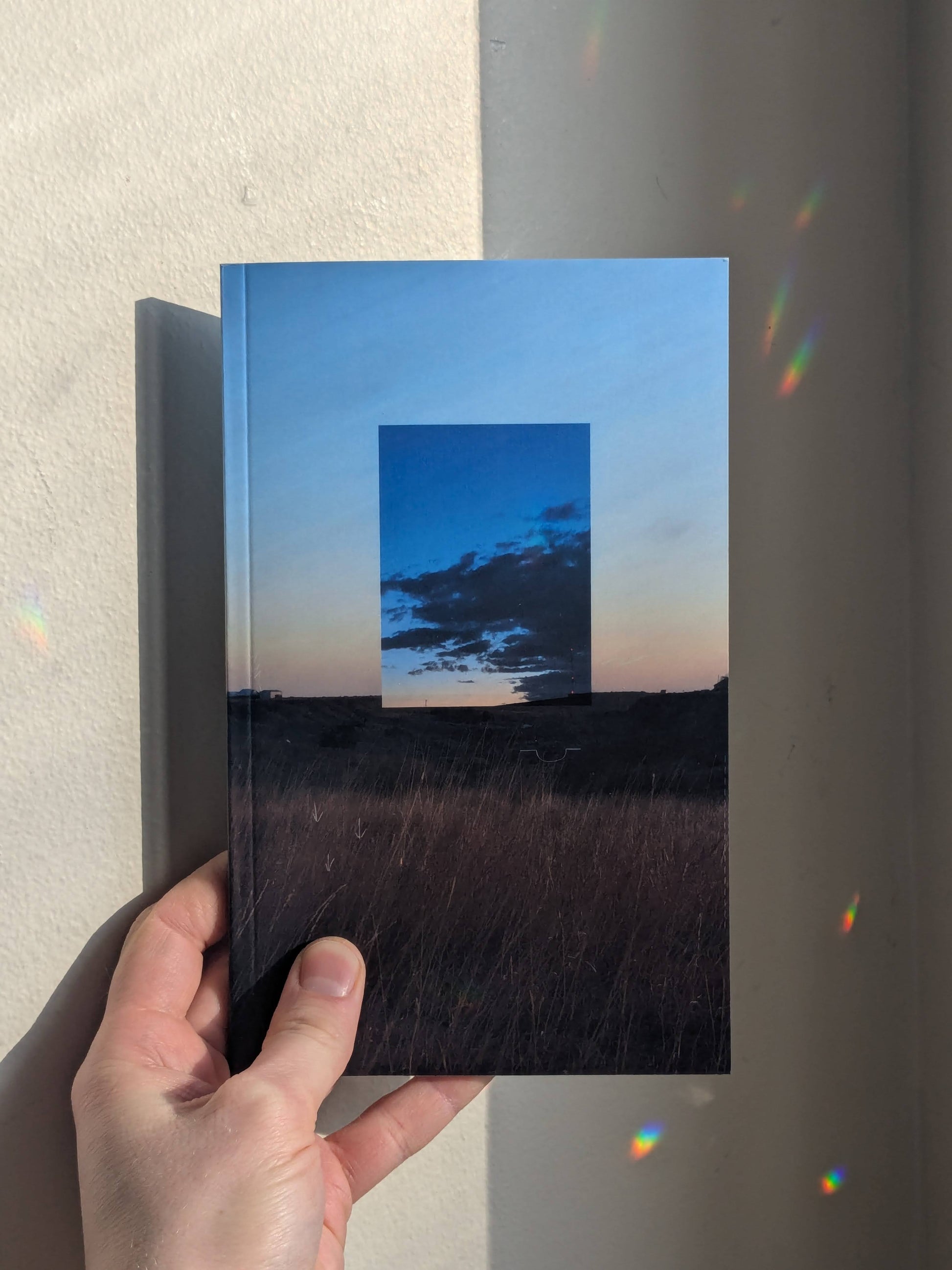 Book featuring a cover of a field, a blue sky, and clouds, held by a hand against a neutral background with refracted light.