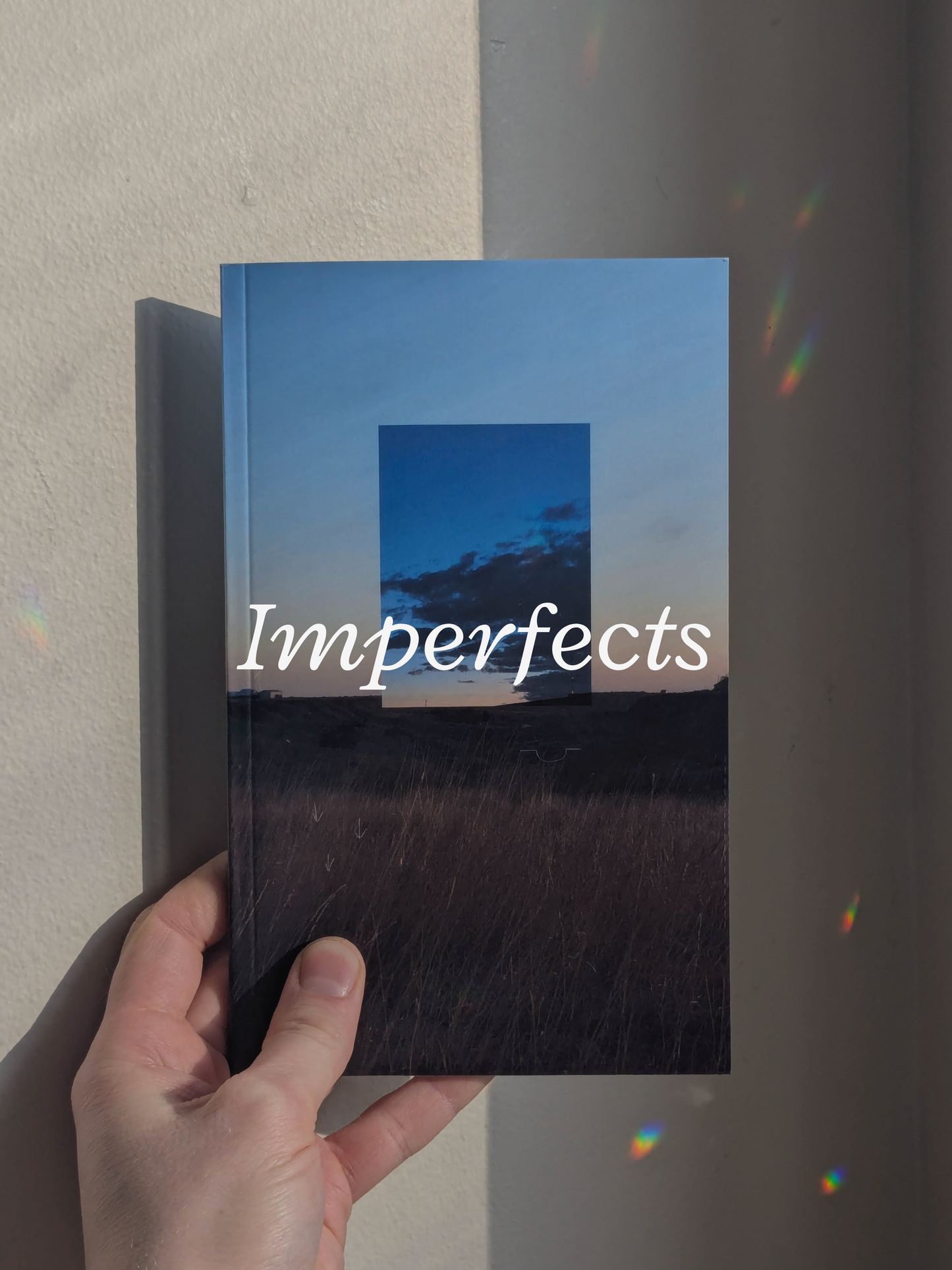 Book featuring a cover of a field, a blue sky, and clouds, held by a hand against a neutral background with refracted light. Across the image reads "Imperfects."